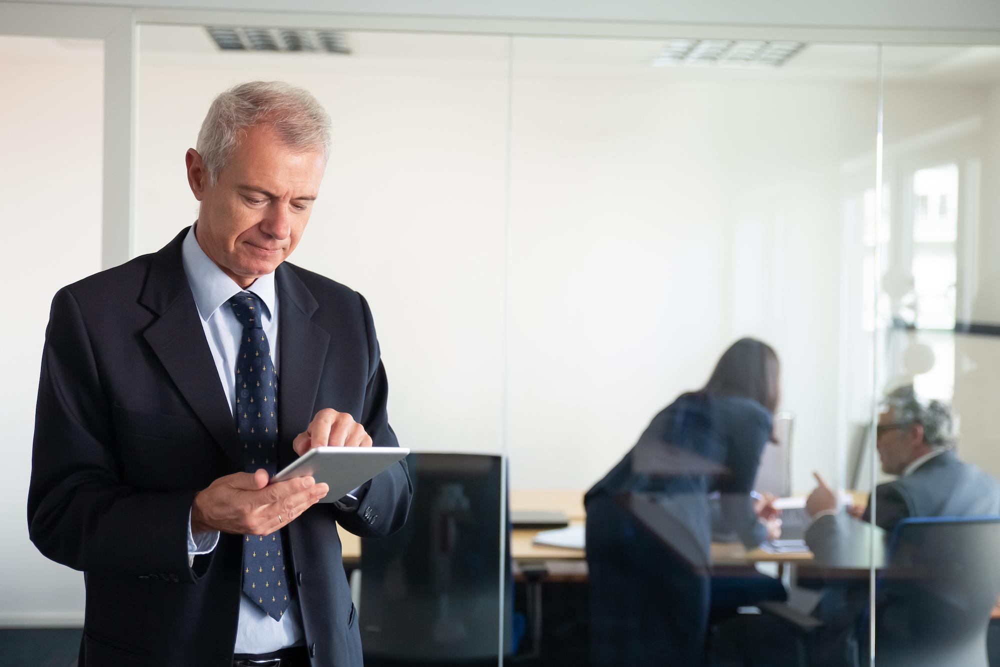 focused-mature-businessman-using-tablet-while-his-colleagues-discussing-project-workplace-glass-wall-copy-space-communication-concept
